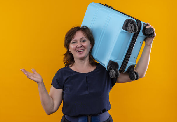 摊开微笑的中年旅行家妇女肩上扛着手提箱 手伸向孤零零的黄色墙壁肩膀微笑中年