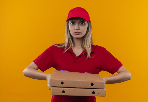 制服严格的年轻送货女孩穿着红色制服和帽子举行比萨饼盒隔离在橙色墙上送货年轻披萨