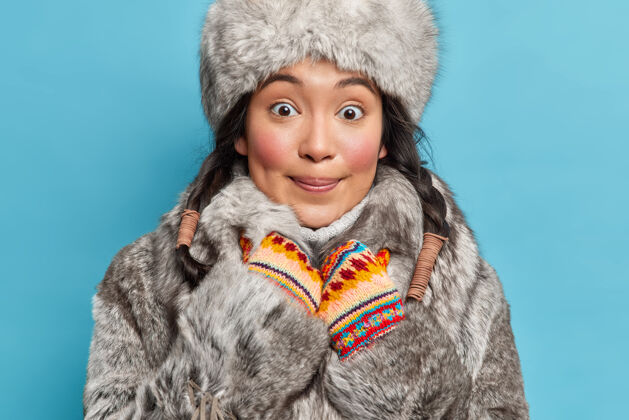 冬天来自阿拉斯加州的快乐女人脸上露出惊讶和喜悦的表情 戴着冬日帽 皮衣和手套 对着蓝色的墙壁摆姿势手套毛衣辫子