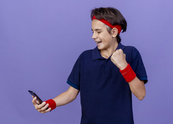 手势快乐的年轻帅气的运动男孩戴着头带和护腕 戴着牙套 拿着手机 在紫色的墙上做着“是”的手势头带牙科手机