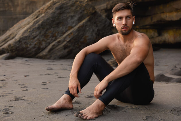 沙滩无忧无虑的男子冠军光着脚坐着 穿着黑色紧身裤 留着胡子射击音频赤脚