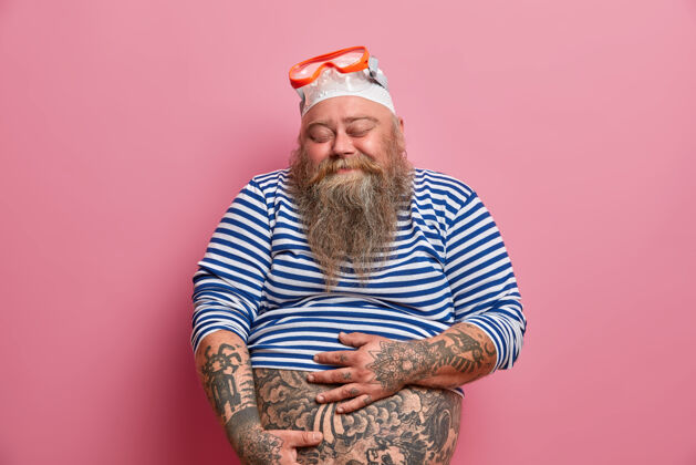 人一张孤立的照片 丰满的胡须男人抱着他那肥嘟嘟的肚子 满意地闭上眼睛 穿着小尺寸的水手衬衫 橡胶泳衣和护目镜 在夏天喜欢浮潜爱好娱乐举行