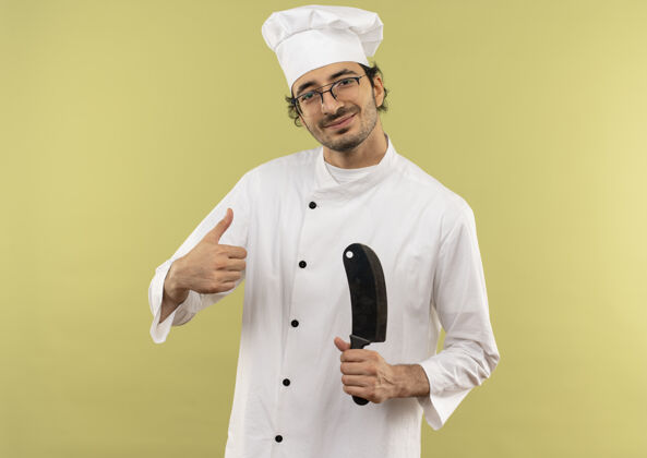 请穿年轻的男厨师穿着厨师制服 戴着眼镜 拿着菜刀 他的大拇指孤立地竖在绿色的墙上手持拇指制服