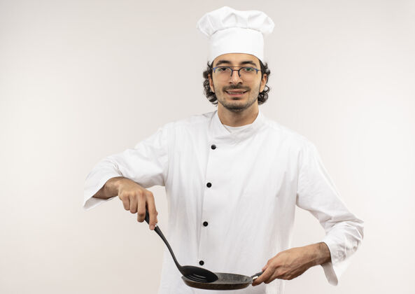 平底锅年轻的男厨师穿着厨师制服 戴着眼镜 手里拿着抹刀和煎锅 隔离在白色的墙上薯条抹刀请穿