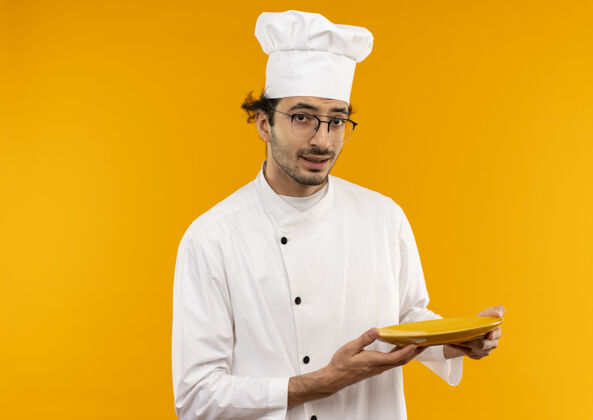 烹饪想着年轻的男厨师穿着厨师制服戴着眼镜把盘子隔离在黄色的墙上佩戴持有男性