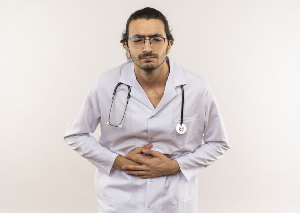 穿生病的年轻男医生戴着眼镜 穿着白色长袍 带着听诊器 在隔离的白色墙壁上抓住疼痛的胃 留着复印空间长袍年轻光学
