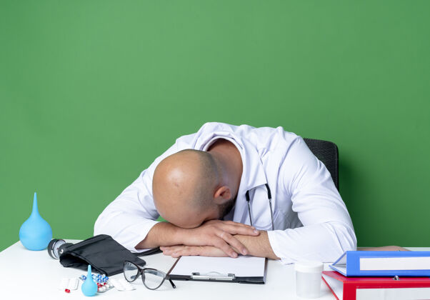 办公桌低着头的年轻男医生穿着医用长袍 听诊器坐在桌子旁 医疗工具被隔离在绿色的墙上听诊器头长袍