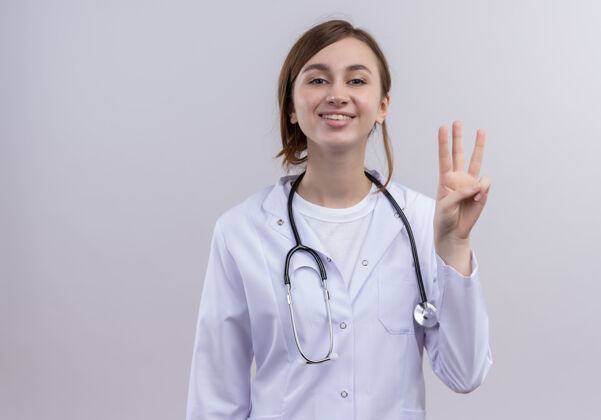 微笑微笑着的年轻女医生穿着医用长袍和听诊器 在隔离的白墙上显示着三个 还有复印空间女人听诊器年轻人