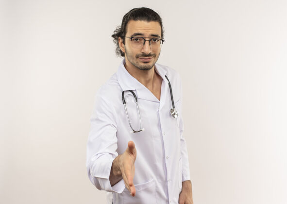 年轻高兴的年轻男医生戴着眼镜 穿着白色长袍 手持听诊器 手伸向隔离的白墙上 留着复印空间男性手持光学