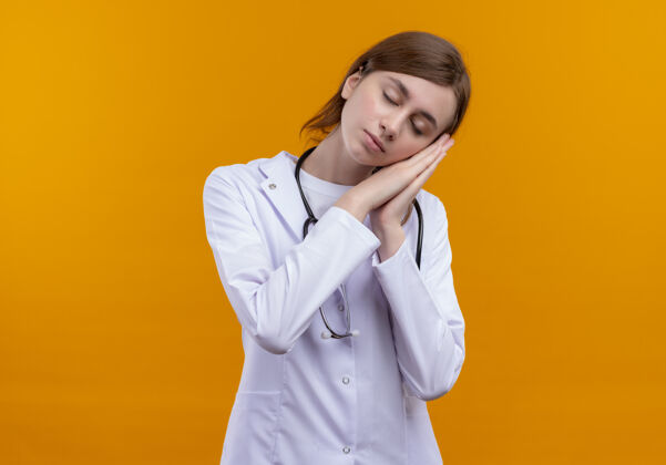 女人疲惫的年轻女医生穿着医用长袍和听诊器在隔离的橙色墙壁上做着睡觉的姿势 还有复印空间长袍疲倦睡眠