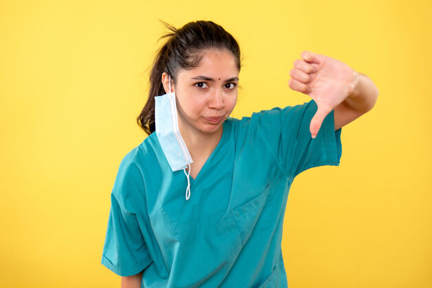 听诊器戴着医用口罩的年轻女性正面图 黄色墙上有拇指朝下的标志制造前面拇指