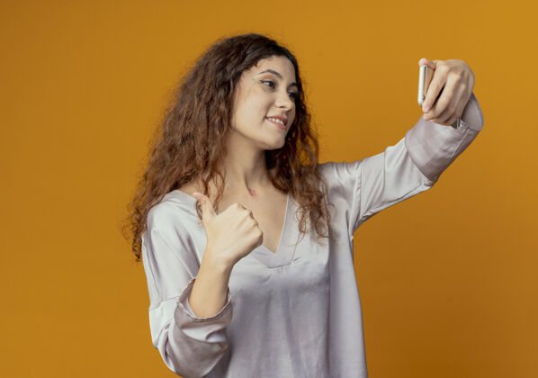 女孩微笑的年轻漂亮女孩自拍她的大拇指孤立在黄色的墙上接受漂亮自拍