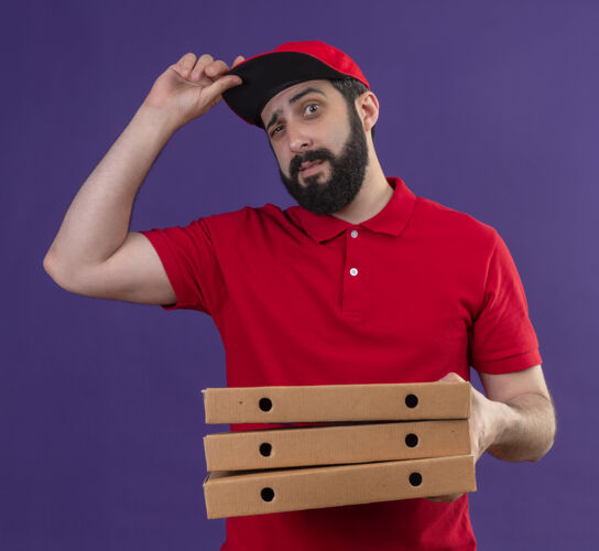 送货自信的年轻英俊的白人送货员穿着红色制服 戴着披萨盒的帽子 帽子上的紫色隔离着年轻帅气帽子