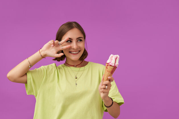 年轻人十几岁的女孩 快乐的 深色头发拿着冰淇淋的女士发出信号 看着右边紫色墙上的复制空间穿着绿色t恤 手镯 戒指和项链黑发项链短发