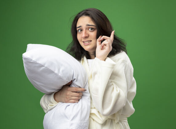 女孩印象深刻的年轻白种人生病的女孩穿着长袍站在侧视图拥抱枕头看着相机指向上绿色背景与复制空间隔离拥抱年轻人长袍