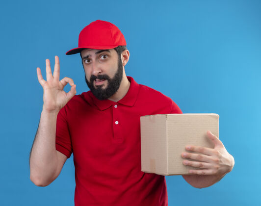 标志自信的年轻英俊的白人送货员穿着红色制服 戴着帽子 拿着纸箱 在蓝色上做着“ok”标志纸箱穿持有