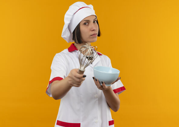 年轻自信的年轻女厨师 身着厨师制服 手拿碗 手拿搅拌器 看起来孤立地站在橙色的空间里厨师拂持有