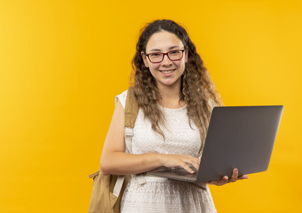 包快乐的微笑着 年轻漂亮的女学生戴着眼镜 背着包拿着笔记本电脑 隔离在黄色的复印空间里穿微笑漂亮