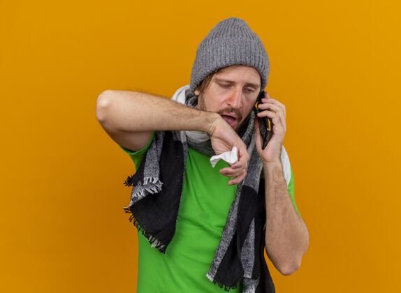 弱瘦弱的年轻英俊的斯拉夫病夫戴着冬天的帽子和围巾打电话拿着餐巾看着笔直的手在空气中隔离在橙色的背景和复制空间年轻电话疾病