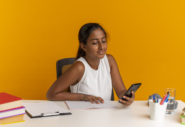 工具高兴的小女生坐在书桌旁 手里拿着学习工具 看着橘色墙上的电话女学生抱着学校
