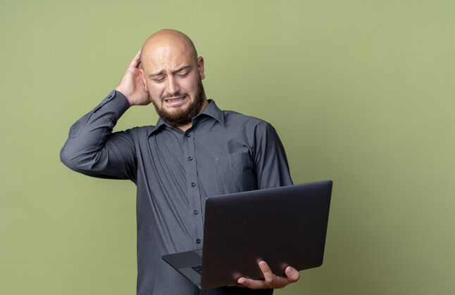 绿色恼怒的年轻秃头呼叫中心男子拿着笔记本电脑 手放在头上 孤立地看着橄榄绿的复印空间年轻秃头笔记本电脑