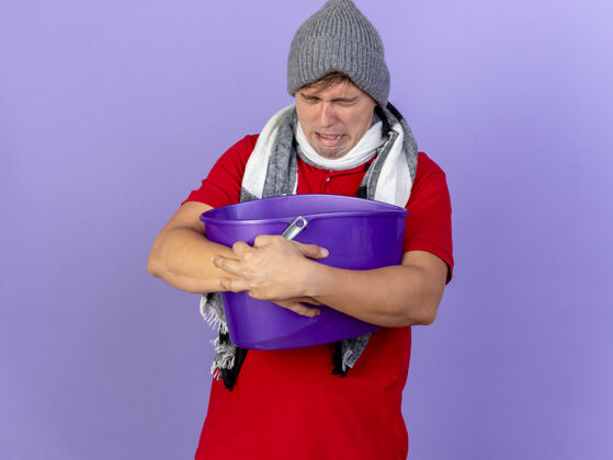 眼睛年轻英俊的金发病男子戴着冬天的帽子和围巾拿着塑料桶恶心闭着眼睛隔离在紫色背景与复制空间疾病塑料水桶