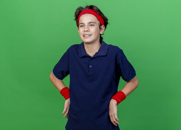 背带令人印象深刻的年轻英俊的运动男孩戴着头带和护腕与牙套保持双手放在腰上看直隔离绿色背景与复制空间年轻腕带运动