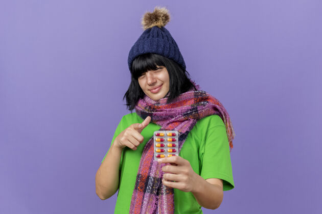 女孩高兴的年轻病高加索女孩戴着冬天的帽子和围巾 拿着一包胶囊 看着和指着相机隔离在紫色背景与复制空间指着胶囊请