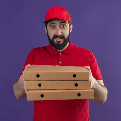 红色印象深刻的年轻英俊的白人送货员穿着红色制服和帽子伸出比萨饼盒孤立的紫色印象帽子盒子