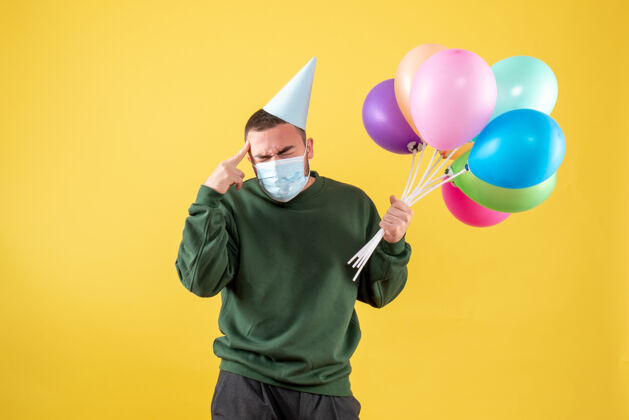 面具正面图：黄色背景上戴着面具手持彩色气球的年轻男性大流行病毒聚会