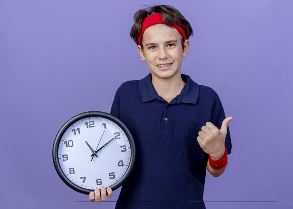 年轻微笑的年轻英俊的运动男孩戴着头带和戴着牙套的腕带看着相机拿着时钟显示拇指向上孤立的紫色背景与复制空间拇指举行头带
