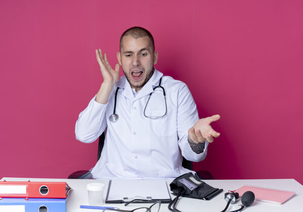 男性恼怒的年轻男医生穿着医用长袍和听诊器坐在办公桌旁 手里拿着工作工具 手举在空中 孤立地看着粉红色的办公桌长袍空气年轻