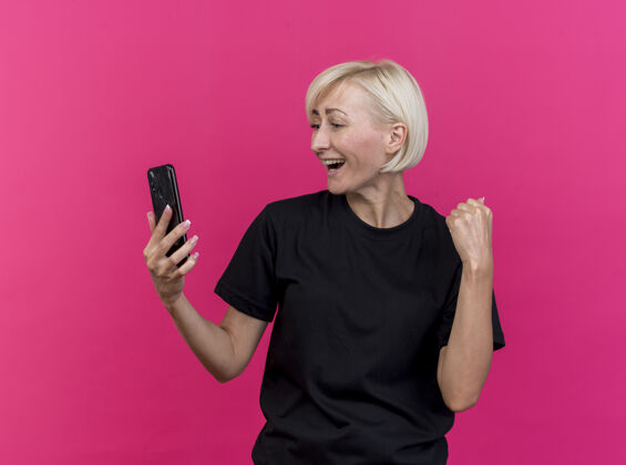 电话快乐的中年金发斯拉夫女人拿着手机 看着手机做着“是”的手势 在深红的背景下与世隔绝金发是的手机
