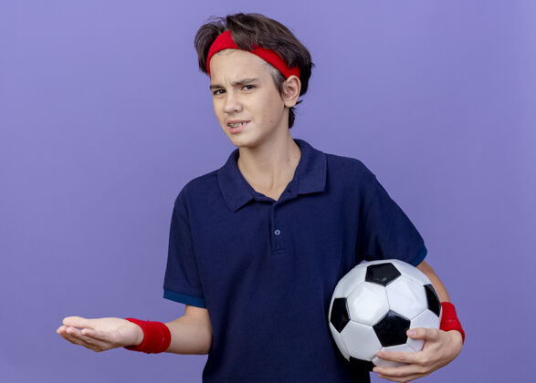 手戴着头带和戴着牙套的腕带的年轻帅气的运动男孩看着拿着足球的摄像机 在紫色背景上显示出孤立的空手男孩球帅气