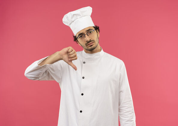 制服悲伤的年轻男厨师穿着厨师制服 戴着眼镜 他的大拇指被隔离在粉红色的墙上男性眼镜穿
