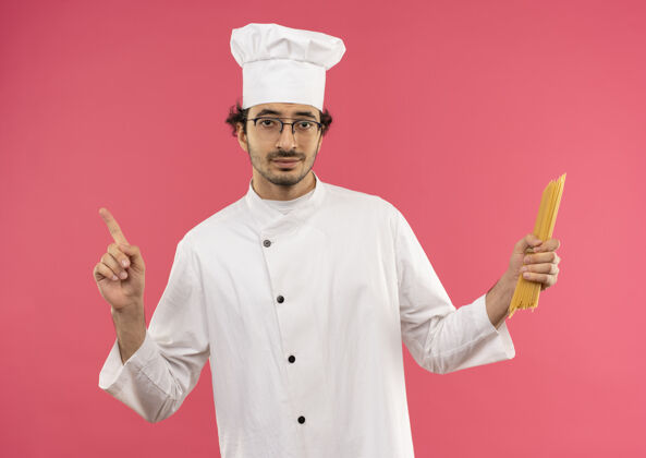 厨师高兴的年轻男厨师穿着厨师制服 戴着眼镜拿着意大利面 指着隔离在粉红色墙上的复制空间年轻点烹饪