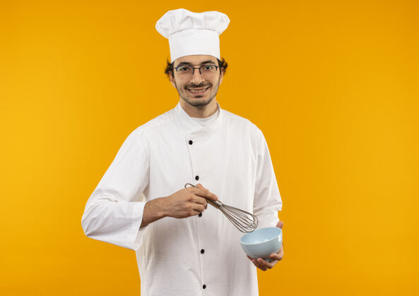 厨师面带微笑的年轻男厨师 身穿厨师制服 戴着眼镜 拿着威士忌和碗 隔离在黄色的墙上烹饪眼镜穿