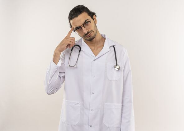 额头年轻的男医生戴着眼镜 穿着白色长袍 听诊器 把手指放在额头上 在隔离的白色墙壁上 留有复印空间思维医生眼镜