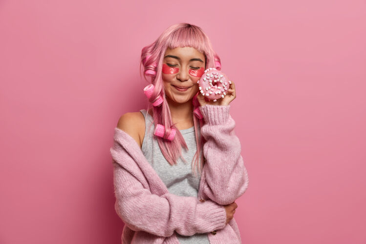 蛋糕一头粉红头发的亚洲年轻女子 闭上眼睛 把美味的甜甜圈贴在脸上 在眼睛下面涂上胶原蛋白 做成卷发纯亚洲人光滑