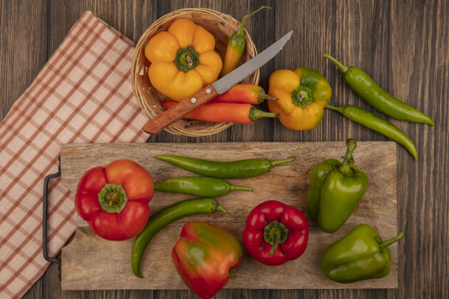 辛辣新鲜的红色和绿色辣椒在木制厨房板上的俯视图 黄色辣椒在木桶上 刀放在木墙上的格子布上查看蔬菜木材