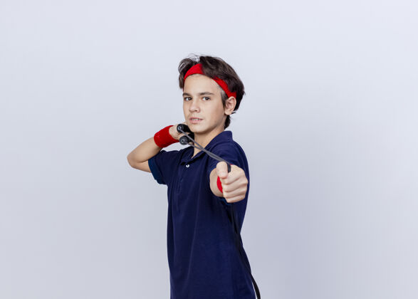 腕带年轻帅气的运动男孩戴着头带和戴着牙套的腕带站在侧视图中拉着白色背景上的跳绳年轻立场复制