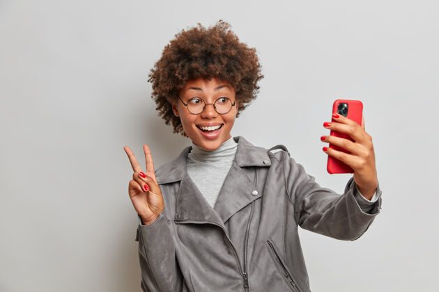 肖像积极无忧无虑的卷发女人在现代智能手机上自拍 做出和平的手势 举起两个手指 有快乐的表情 享受业余时间 穿着时髦的夹克眼镜时尚乐观