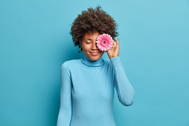 封面年轻快乐的黑皮肤女人用非洲菊遮住眼睛 从地里采花 去装饰室 穿着休闲的蓝色高领毛衣蓝色发型粉色