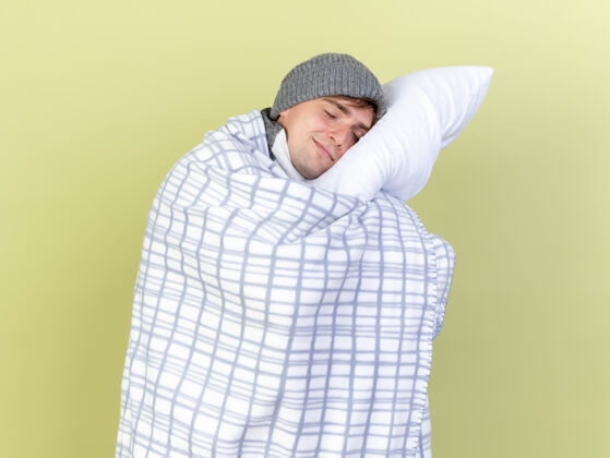 头高兴的年轻英俊的金发病男子戴着冬天的帽子和围巾在格子布包裹抱着枕头把头放在它与橄榄绿的背景隔离眼睛封闭穿包裹