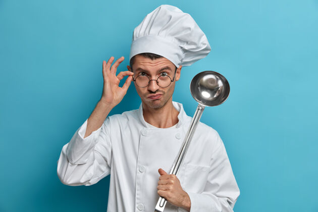 认真厨师长在商业厨房摆姿势 透过眼镜严肃地看着 拿着勺子 准备饭菜 准备做饭 听取建议自信咖啡馆器皿