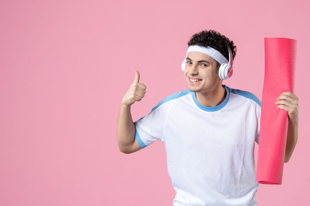 水平正面图穿着运动服的年轻男性 粉色墙上有瑜伽垫成人竖起大拇指运动