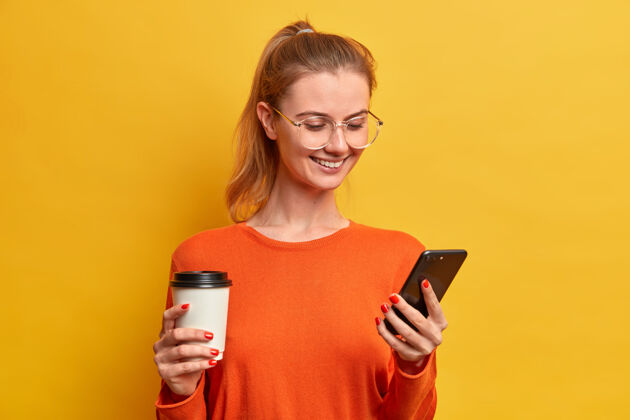 黄色快乐美丽的千禧一代女孩下载新的移动应用程序 喝咖啡从纸杯 有愉快的微笑 短信聊天 戴眼镜 梳头在马尾 上网外卖冲浪骄傲