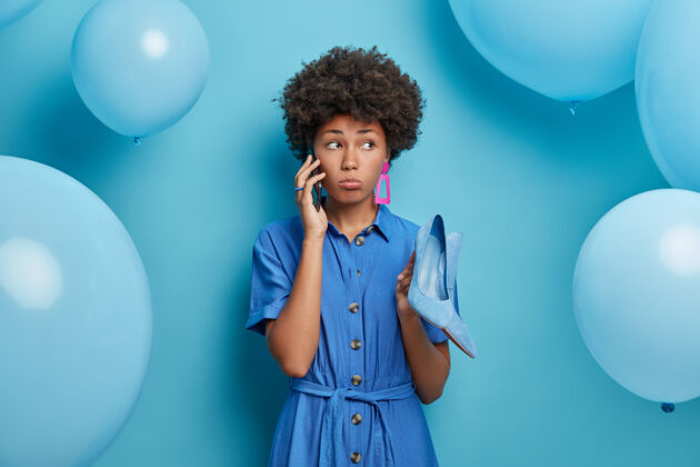 牙齿一切都是蓝色的悲伤失望的非裔美国妇女对重新安排的聚会不满意 通过智能手机给最好的朋友打电话 穿着时髦的高跟鞋 到处放着充气气球服饰女性选择