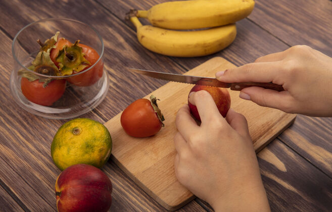 板俯视图中的女性手切割一个新鲜的桃子在一个木制的厨房板上用刀与橘子和香蕉隔离在木墙上女性农业新鲜