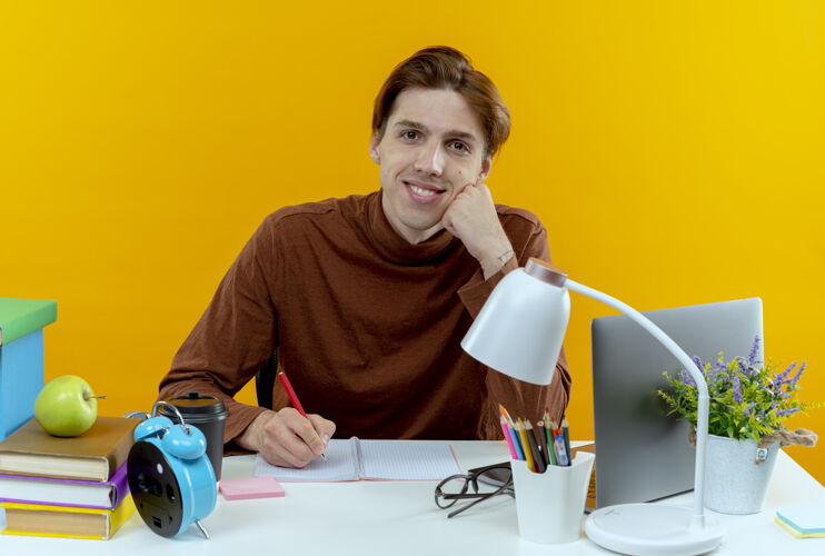 年轻微笑的年轻学生男孩坐在书桌旁 手里拿着学习工具 在黄色的笔记本上写着什么黄色学校男孩